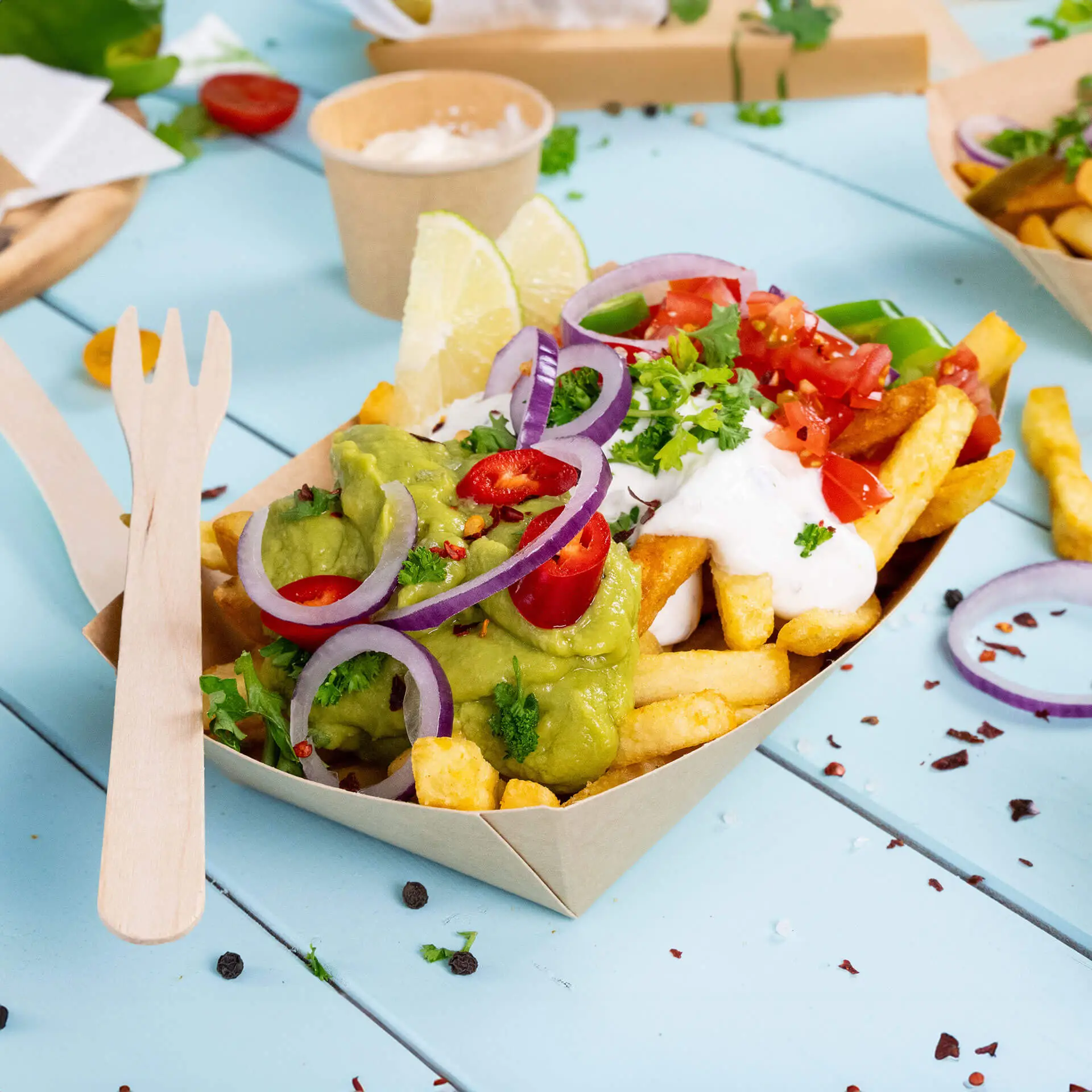 A portion of fries topped with guacamole, diced tomatoes, sour cream, and red onion served in a brown cardboard snack tray.